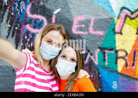 Buone amici femminili che prendono selfie attraverso lo smartphone mentre gesturing contro la parete graffiti Foto Stock