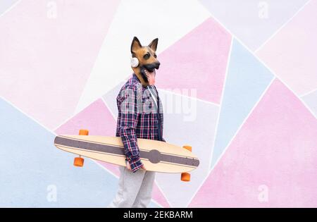 Donna che indossa una maschera per cani che tiene lo skateboard mentre si trova contro il muro Foto Stock