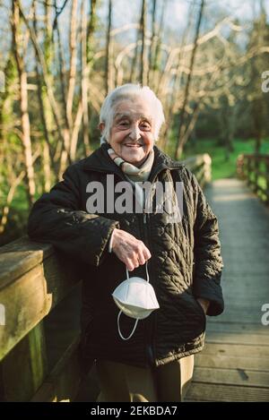 Donna anziana sorridente sul passerella che tiene la maschera protettiva del viso durante COVID-19 Foto Stock