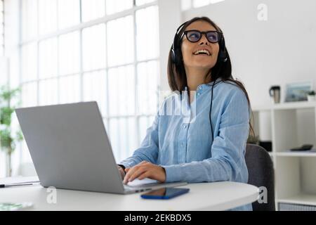 Donna d'affari sorridente che indossa cuffie con microfono che si guarda lontano mentre si è seduti computer portatile in ufficio Foto Stock