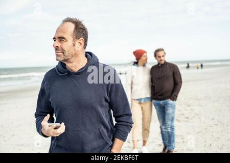 Ritratto dell'uomo in piedi sulla spiaggia sabbiosa con smartphone in mano con giovane coppia in background Foto Stock