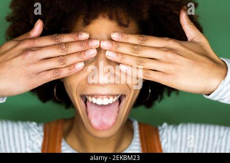 Giovane donna allegra che si stacca la lingua mentre copre gli occhi contro parete verde Foto Stock