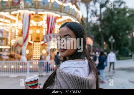 Bella giovane donna che guarda sopra la spalla mentre si sta in piedi contro illuminato carosello Foto Stock