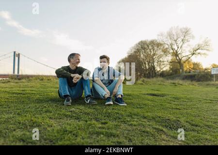 Felice padre e figlio che parlano tra loro mentre si siedono su erba contro cielo Foto Stock