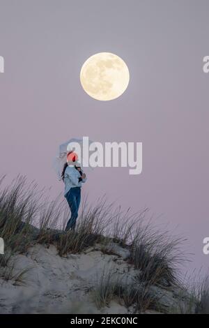 Giovane donna in cappello a maglia arancione che tiene l'ombrello mentre si è in piedi sulla duna di sabbia durante il tramonto Foto Stock