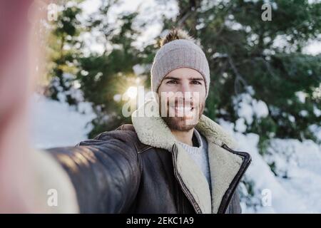 Felice giovane uomo che prende selfie contro gli alberi in tempo freddo Foto Stock