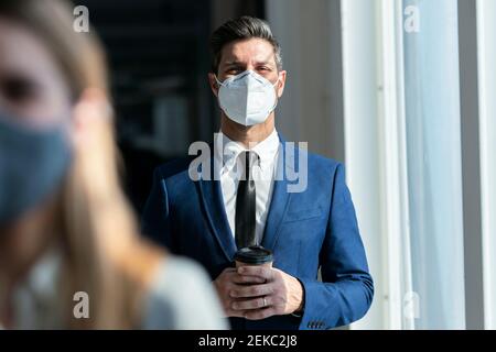 Uomo d'affari che indossa una maschera protettiva che tiene il caffè mentre si sta in piedi ufficio Foto Stock