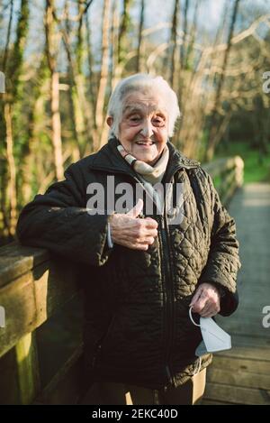 Donna anziana sorridente che mostra i pollici mentre si sta in piedi sul ponte pedonale Durante COVID-19 Foto Stock