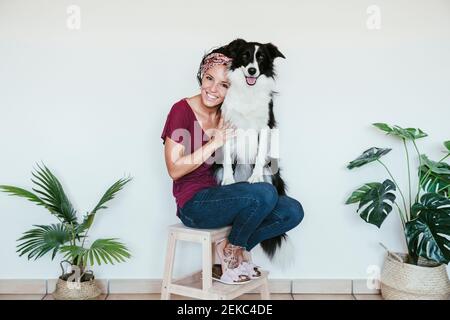 Donna sorridente che abbraccia Border Collie mentre si siede su sgabello in mezzo piante in vaso a casa Foto Stock