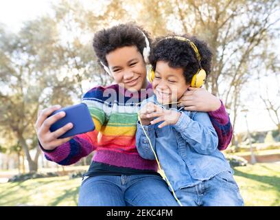 Sibilo indossando cuffie sorridenti mentre si prende selfie attraverso il telefono cellulare mentre si siede al parco Foto Stock