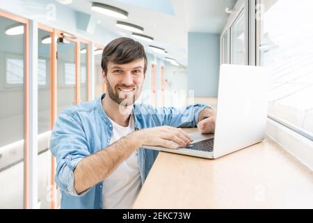 Felice professionista maschile creativo con computer portatile in piedi in corridoio a. luogo di lavoro Foto Stock