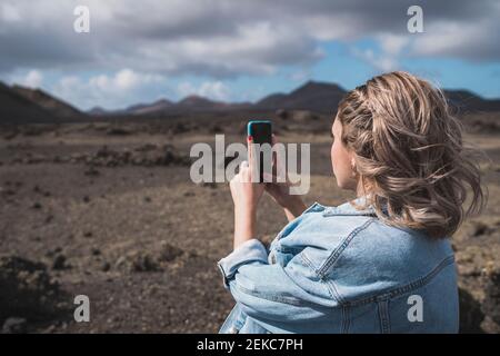 Giovane donna che scatta foto con il cellulare mentre si trova a Volcano El Cuervo, Lanzarote, Spagna Foto Stock