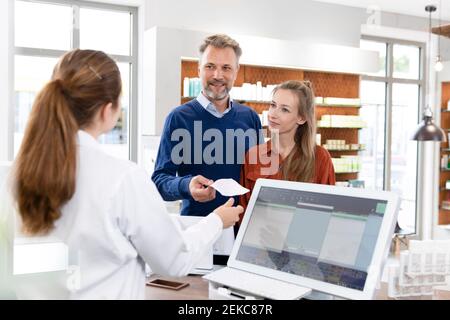 Cliente di sesso maschile che dà l'elenco di prescrizione al farmacista femminile in farmacia pagamento Foto Stock