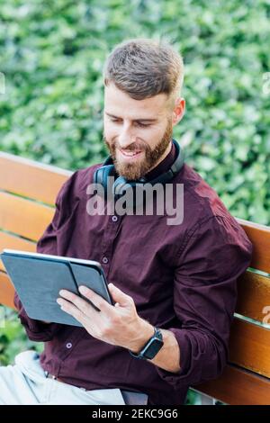 Uomo d'affari sorridente con le cuffie che usano il tablet digitale mentre si è seduti banco Foto Stock