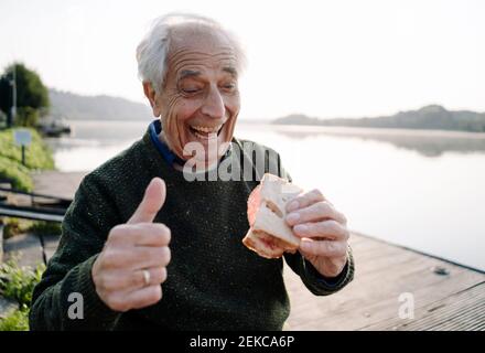 Uomo felice che mostra i pollici mentre mangiano panino seduto sopra molo Foto Stock