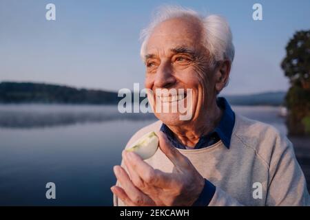 Uomo sorridente che mangia la mela mentre si alza contro il lago Foto Stock