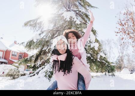 Madre sorridente che fa un giro in piggyback alla figlia con la mano sollevata a terra durante l'inverno Foto Stock