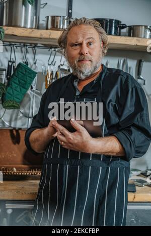 Chef maturo che indossa un grembiule utilizzando un tablet digitale mentre si è in piedi cucina Foto Stock