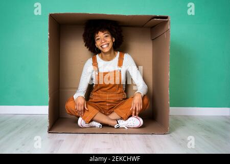 Felice giovane donna seduta con gambe incrociate in scatola di cartone contro il muro verde nella nuova casa Foto Stock