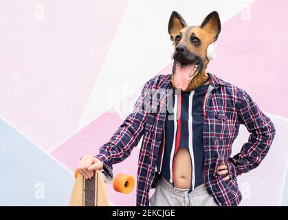 Donna che indossa una maschera per cani e cuffie che tengono lo skateboard mentre si è in piedi contro muro Foto Stock