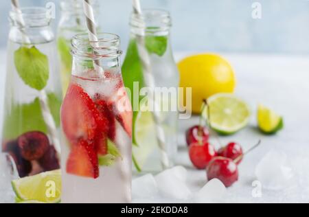 Tante bottiglie con rinfrescante limonata estiva con lime, fragole, ciliegie, cetrioli e ghiaccio su fondo grigio di cemento Foto Stock