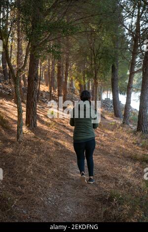 Donna caucasica con giacca verde che cammina su un sentiero tra pini e un lago in una giornata di sole Foto Stock