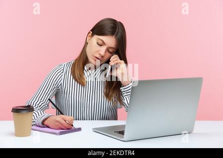 Segretaria diligente in camicia a righe che riceve le chiamate sul telefono e fa le note, che scrive le informazioni, sedendosi al posto di lavoro con il laptop. Prigioniero interno Foto Stock