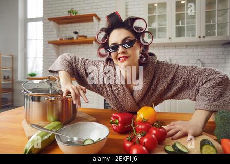 Divertente e bella donna pazza in arricciacapelli e accappatoio preparando la colazione in cucina. Foto Stock