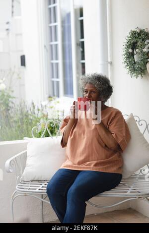 Donna anziana afro-americana premurosa che tiene una tazza di caffè mentre si siede sul portico della casa Foto Stock