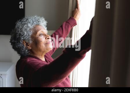 Donna anziana afro-americana premurosa che apre tende a finestre a casa Foto Stock