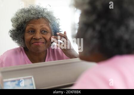 Donna anziana afroamericana sorridente mentre la tocca mentre guardando nello specchio a casa Foto Stock