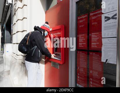 CALDES DE MONTBUI, SPAGNA - 31 GENNAIO 2021: Giovane uomo che indossa un cappello e maschera protettiva, sta utilizzando l'atm della banca Santander in Catalonia Spain du Foto Stock