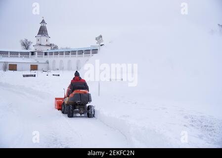 Russia, regione di Mosca, febbraio 2021. UN uomo vestito di calore su un aratro di neve rimuove la neve sul territorio del monastero in una giornata gelida. Foto Stock