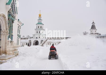 Russia, regione di Mosca, febbraio 2021. UN uomo vestito di calore su un aratro di neve rimuove la neve sul territorio del monastero in una giornata gelida. Foto Stock