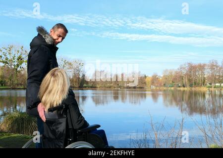 Concetto di persona disabile. Una donna in sedia a rotelle con un uomo in piedi accanto a lei, all'esterno nella natura di fronte ad un lago Foto Stock