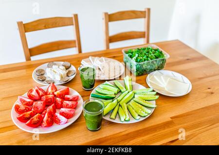 Tavolo in legno con impostazione di sano pranzo vegetariano vegano succo di verdure verdi, formaggio, insalata cetrioli pomodori e nessuno in casa Foto Stock