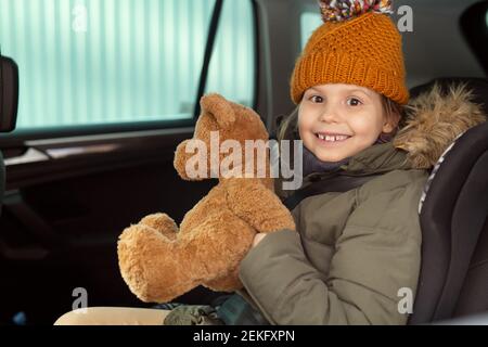 Allegra ragazza carina in winterwear caldo che tiene marrone morbido teddybear seduto sul sedile posteriore dell'auto di fronte fotocamera e ti guarda Foto Stock