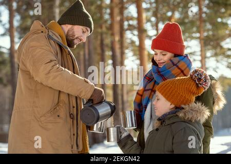 Felice giovane uomo che versa il tè caldo in tazze metalliche di sua moglie e sua figlia mentre si levano in piedi davanti alla macchina fotografica contro alberi di pino Foto Stock