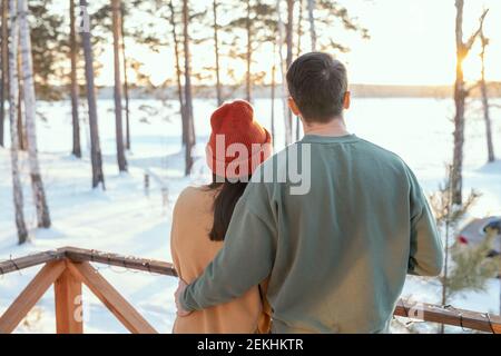Vista posteriore del giovane affettuoso uomo in maglione abbracciando il suo moglie mentre entrambi sono in piedi davanti alla macchina fotografica sul patio della loro casa di campagna Foto Stock
