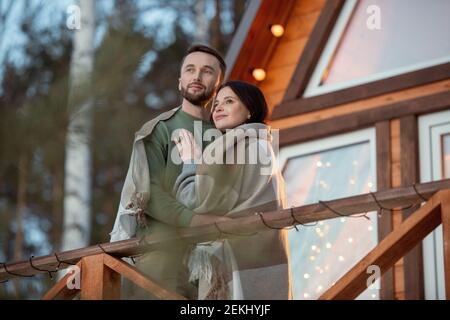 Giovane coppia affettuosa avvolta in plaid in piedi contro casa di campagna sullo sfondo di alberi nella foresta e guardando alba Foto Stock