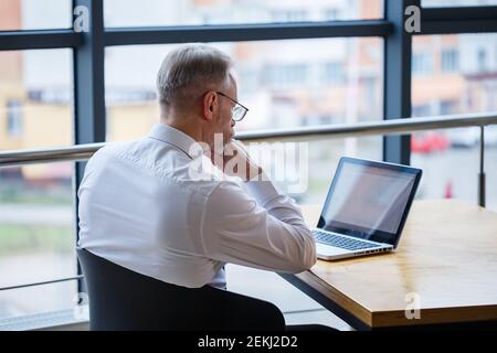 Il freelancer maschile sta lavorando in un caffè su un nuovo progetto di business. Siede in una grande finestra al tavolo. Guarda lo schermo di un computer portatile con una tazza di caffè Foto Stock