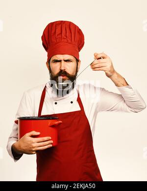Cucinare con un rigoroso volto in Borgogna è uniforme casseruola con siviera. Cucina professionale concetto. Chef zuppa di gusti o di frutta sciroppata. Uomo con barba detiene pot rosso su sfondo bianco. Foto Stock
