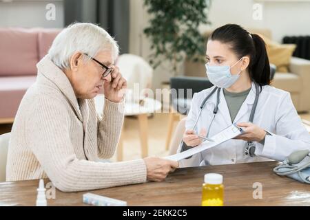 Uomo anziano malato che guarda attraverso un documento medico tenuto da giovani donna medico seduto da tavolo davanti a lui durante consulenza a domicilio Foto Stock
