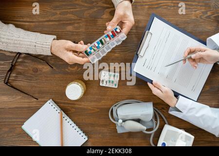 Panoramica delle mani di un uomo anziano malato che tiene il contenitore con pillole sopra la tabella durante la consultazione medica con il medico che si riempie documento Foto Stock