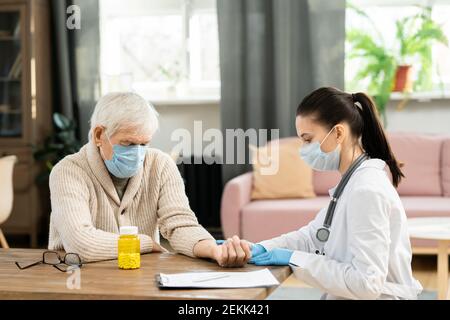 Giovane donna praticante generale in contatto con la maschera protettiva e il bianccoat polso di un paziente malato di sesso maschile senior durante il controllo medico Foto Stock