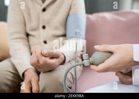 Mano di giovane medico che preme pompa del tonometro mentre misurazione della pressione sanguigna di un anziano malato seduto di fronte di lei Foto Stock