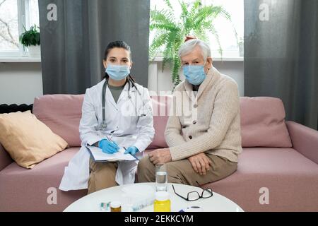 Malato paziente anziano e giovane medico generico in maschere protettive sedersi sul divano uno accanto all'altro davanti a. fotocamera in soggiorno Foto Stock