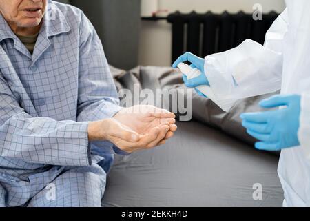 Guanti mani di giovane medico in tute protettive spray disinfettante su quelli dell'anziano malato in pigiama blu seduto davanti a lei Foto Stock