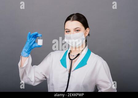 Grave medico femminile in una maschera medica che contiene siringa e. vaccino isolato su grigio Foto Stock