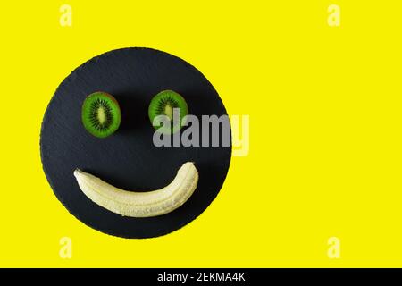 Metà Kiwi e una banana sbucciata su vassoio nero e. sfondo giallo Foto Stock